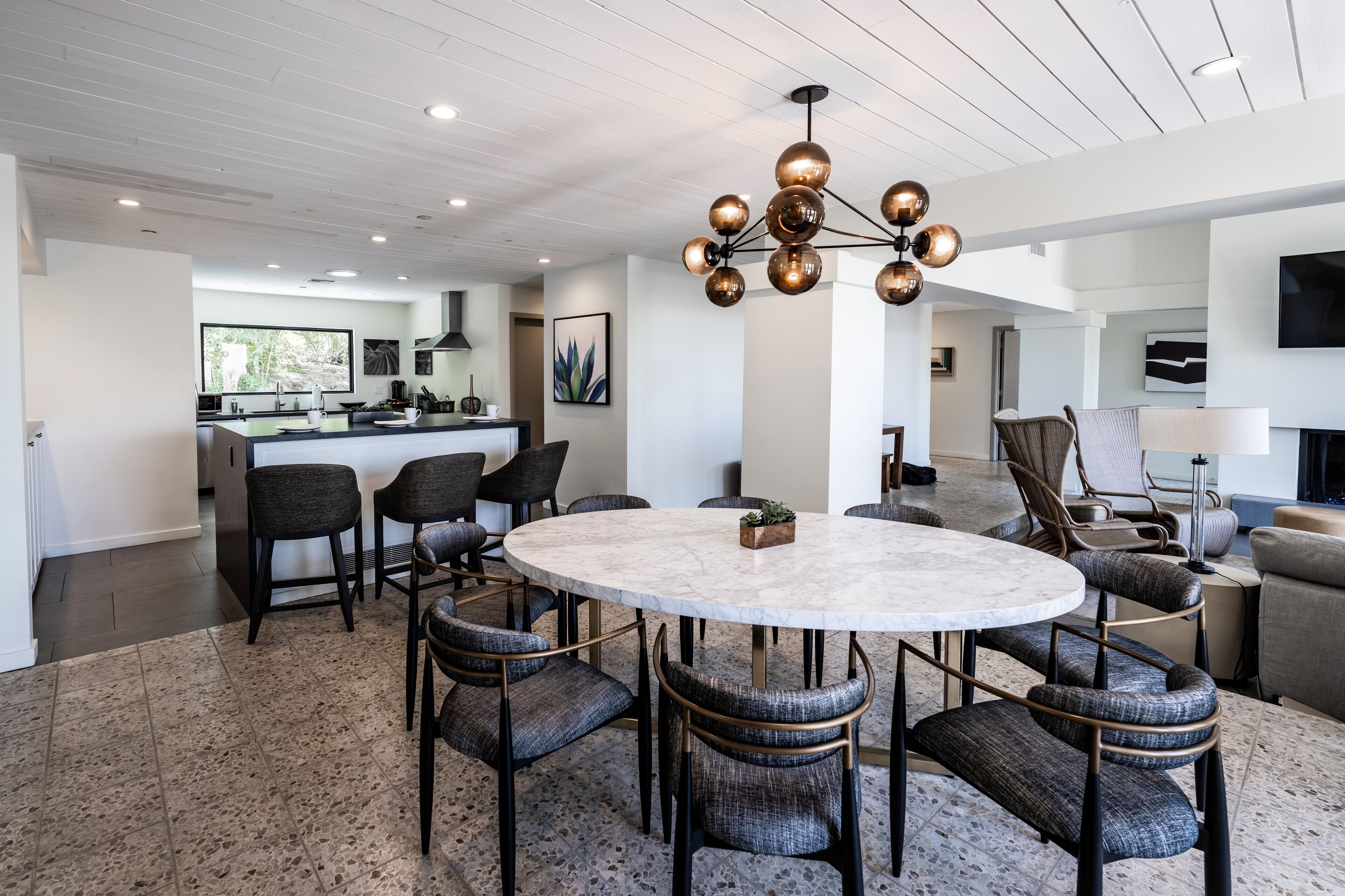 Una sala de estar moderna con una mesa redonda de mármol, sillas tapizadas oscuras, un candelabro central y una cocina al fondo.