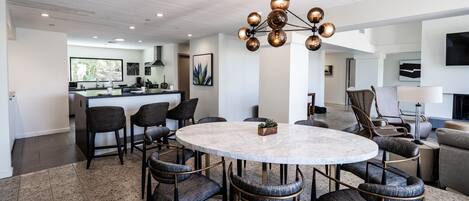 A modern living room with a round marble table, dark upholstered chairs, a central chandelier, and a kitchen in the background.