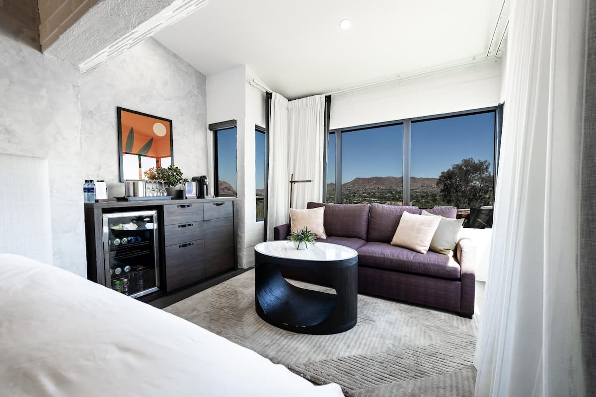 A modern hotel room with a large bed, a round coffee table, a sofa, a minibar, and a large window offering a view of the landscape.