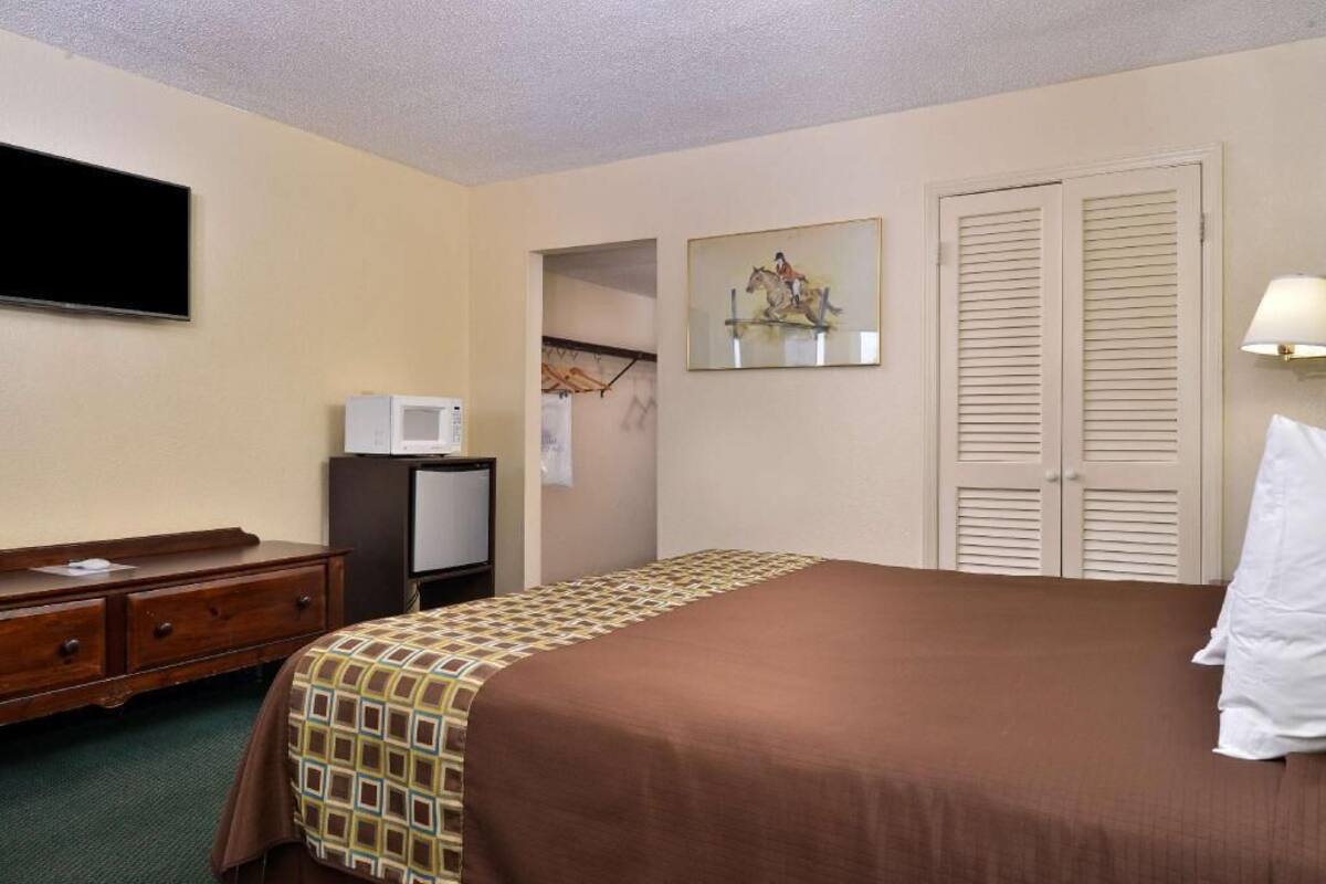Ein Hotelzimmer mit Bett, Fernseher, Schreibtisch mit Computer, Kleiderschrank und einem Bild an der Wand.