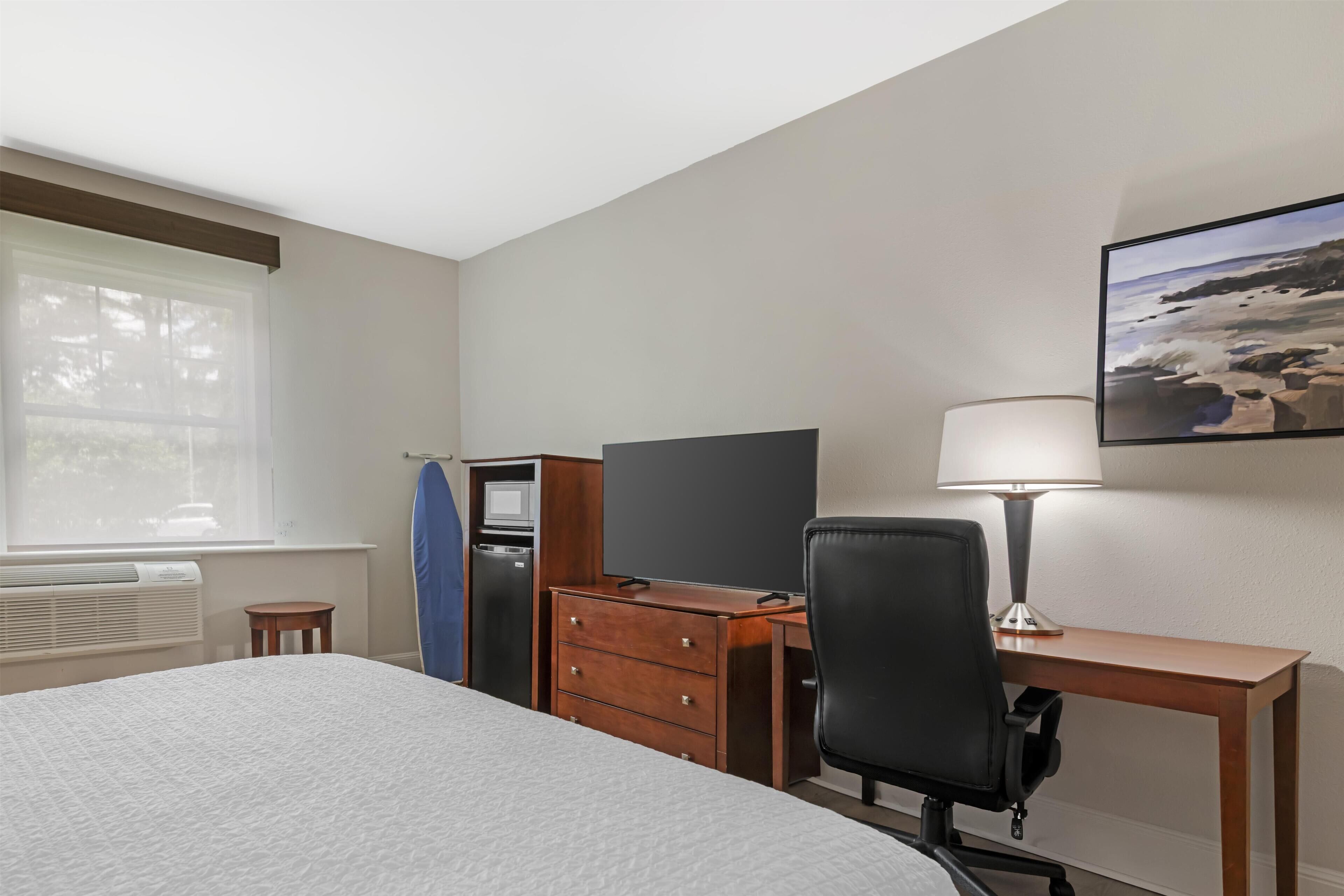 A hotel room with a bed, desk, chair, dresser, and a painting of a beach scene.