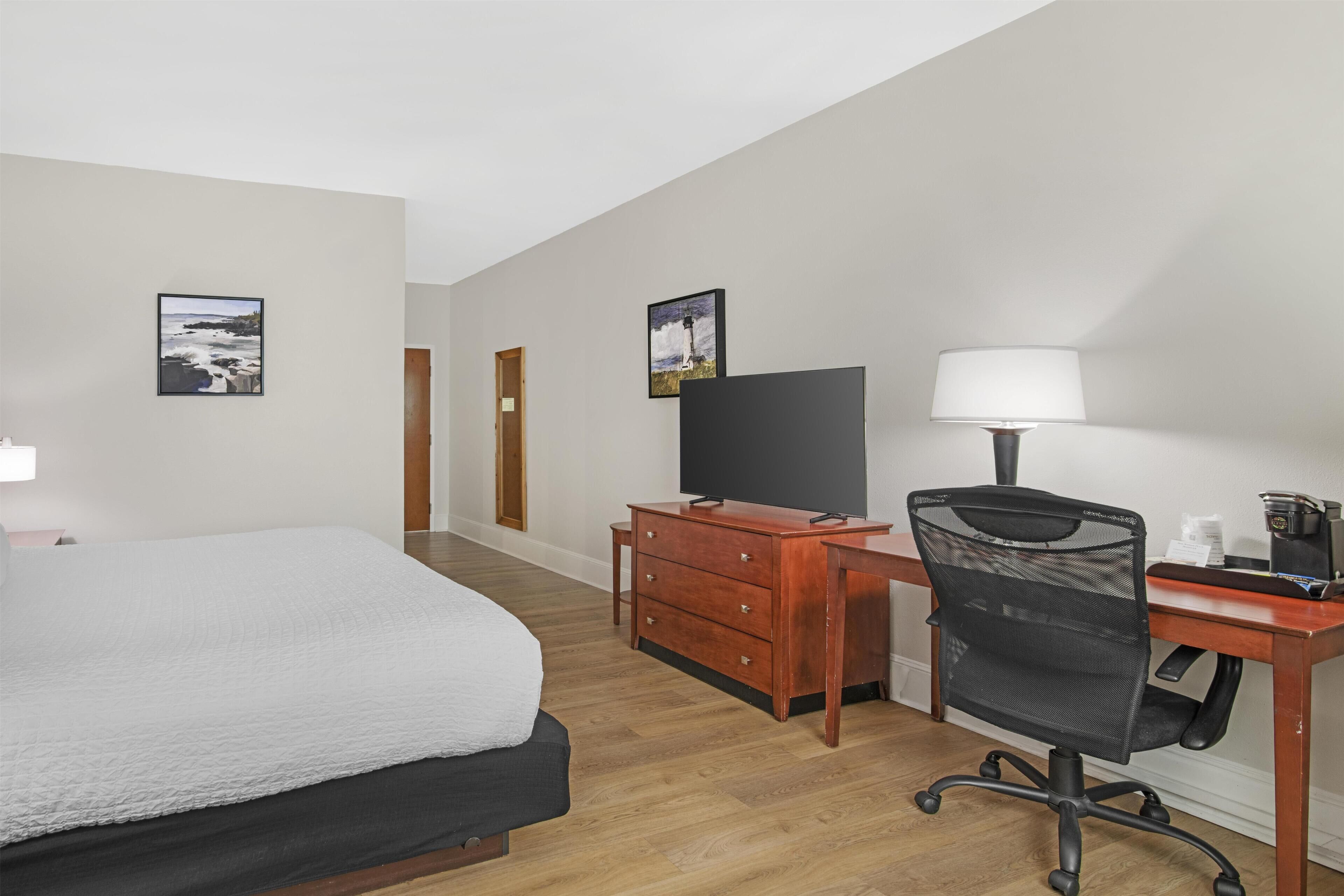 A hotel room with a bed, a desk with a chair, a television, a lamp, and two framed pictures on the wall.