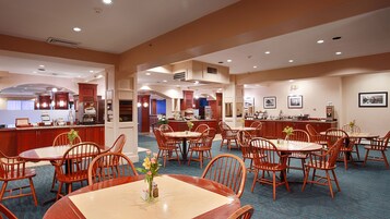 Un comedor de hotel con varias mesas y sillas redondas, una cocina al fondo y una zona de bar.