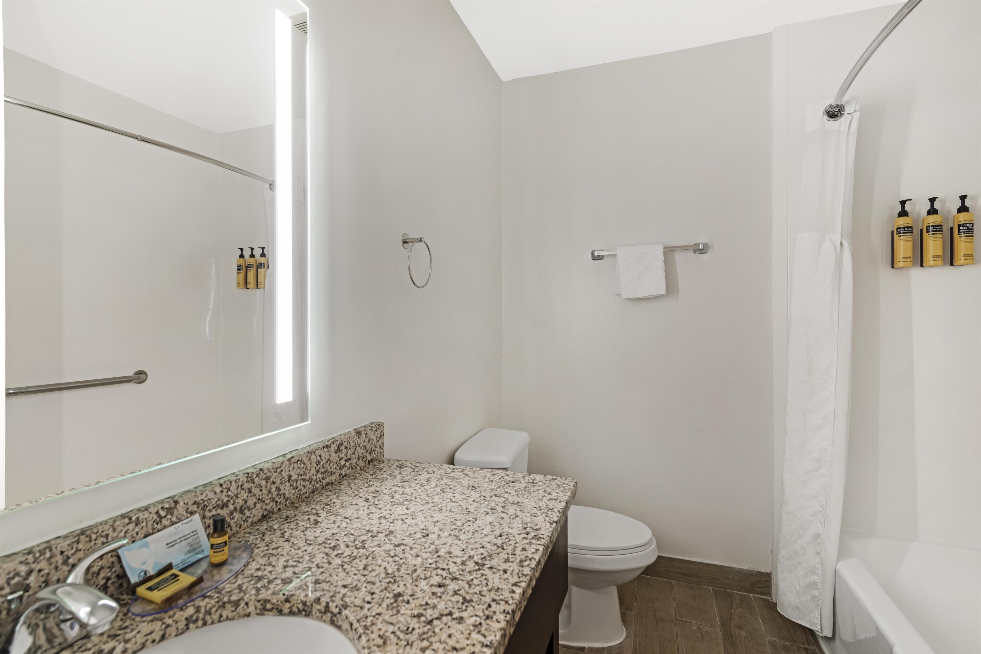 A bathroom with a granite countertop, a white toilet, a shower with a white curtain, and a mirror.