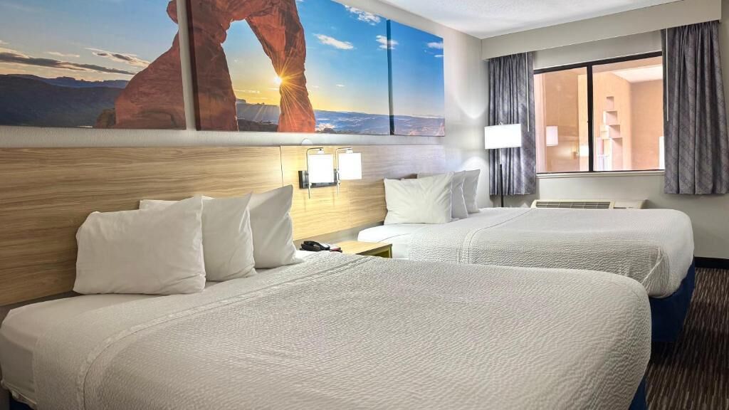 A hotel room with two beds, a large window with curtains, a wall-mounted lamp, and a painting of a rock formation.