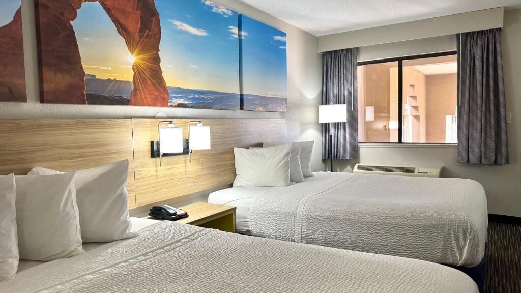 A hotel room with two beds, a large window with curtains, a wall-mounted TV, and a painting of a landscape.