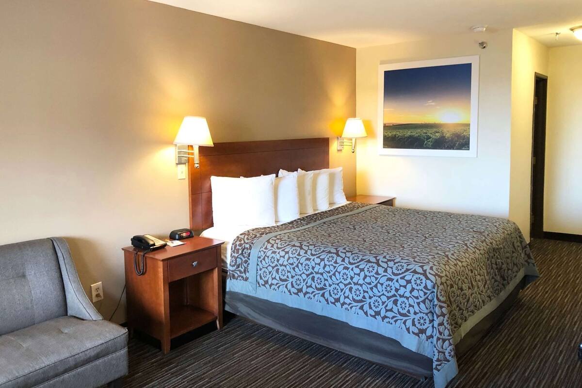 A hotel room with a bed, two bedside lamps, a nightstand with a phone, a chair, and a framed picture on the wall.