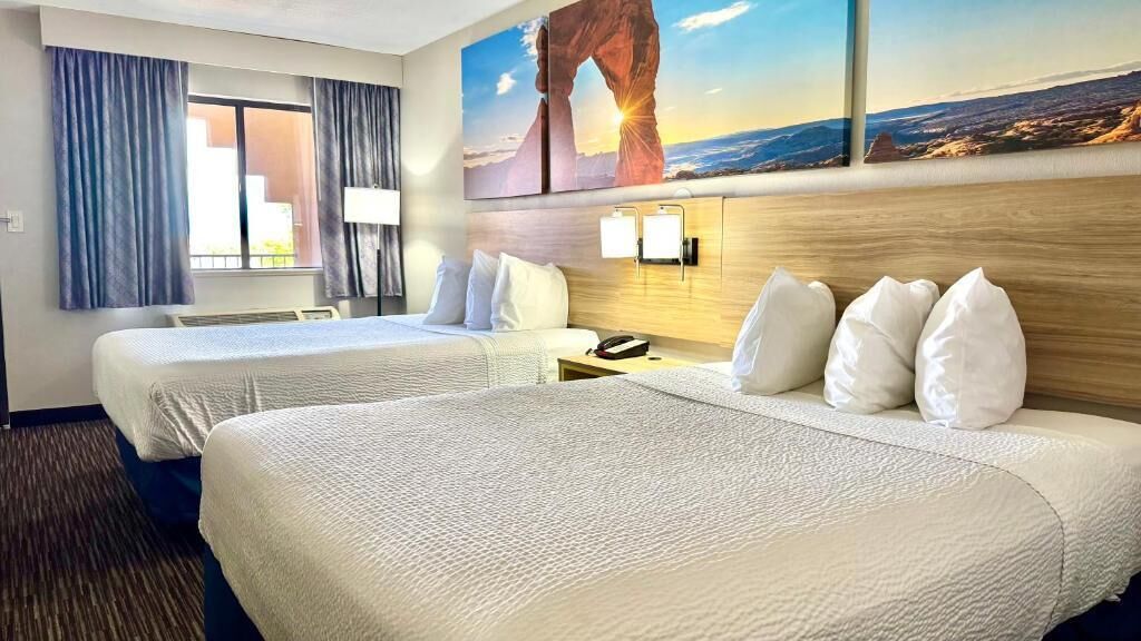 A hotel room with two beds, a nightstand, a lamp, and a painting of a landscape.