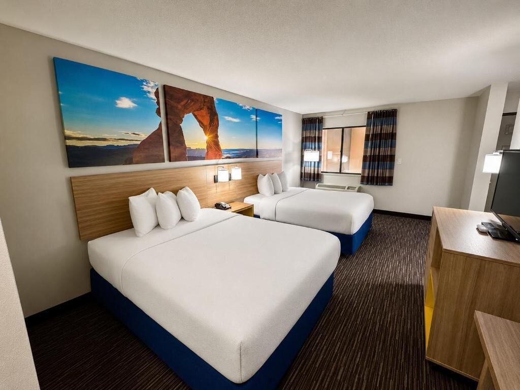 Ein Hotelzimmer mit zwei Betten, einem Schreibtisch, einem Stuhl, einem Fernseher und einem großen Fenster mit Vorhängen.