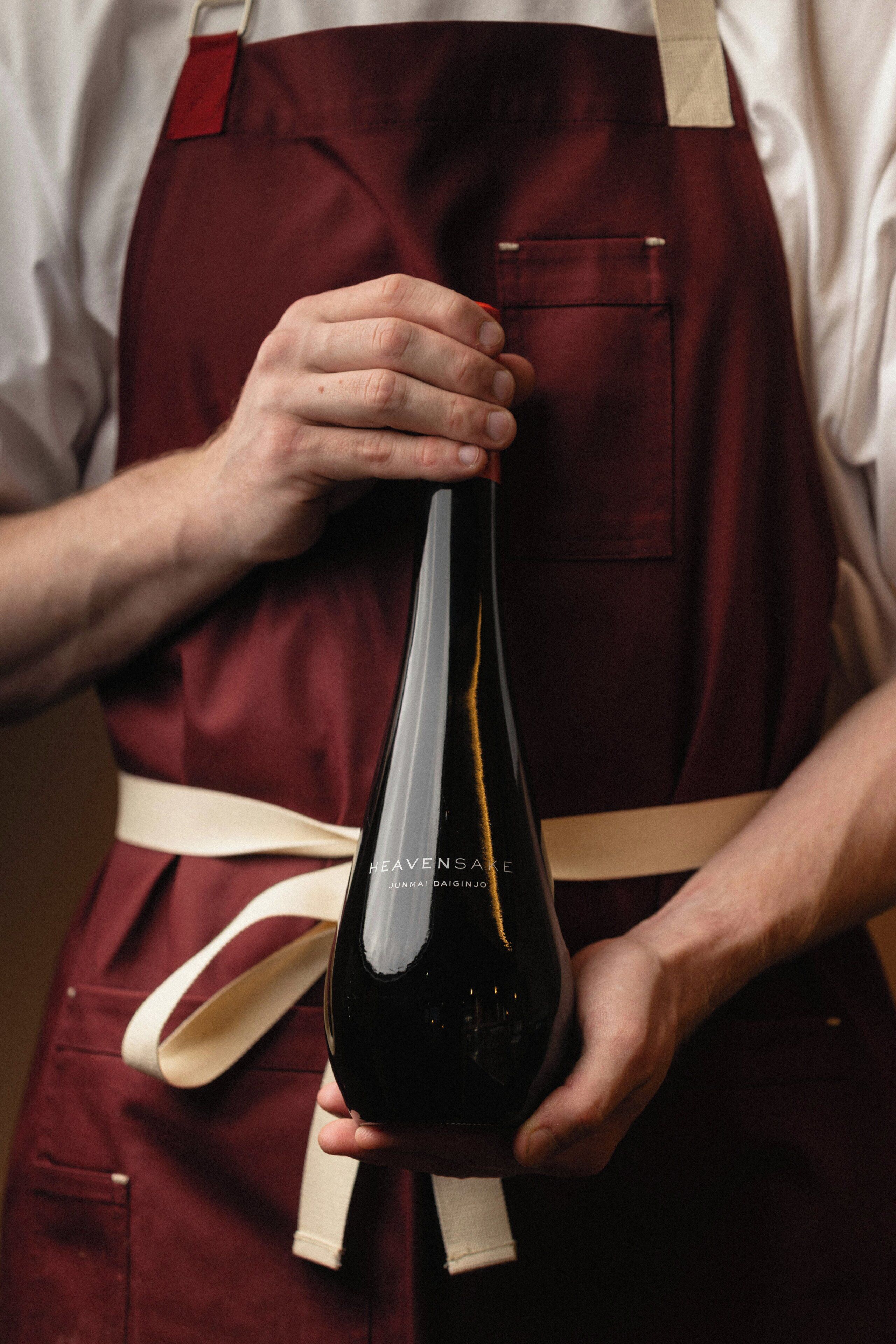A person holding a wine bottle with a label that reads 'HEAVENSAKE JUNMAI DAIGINJO.'