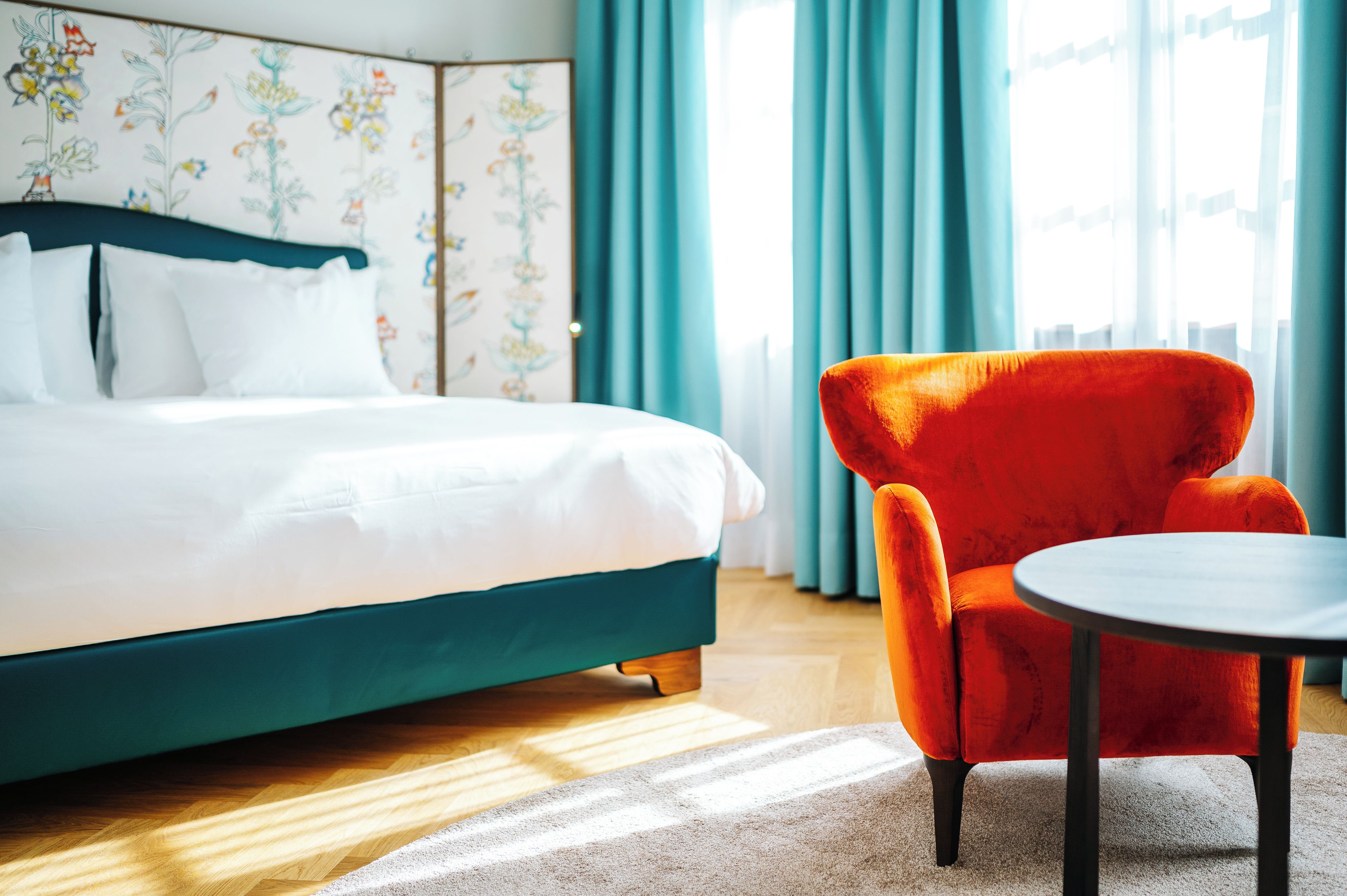 Un lit avec une literie blanche et une tête de lit turquoise, un fauteuil rouge et une petite table ronde dans une pièce dont les murs sont recouverts d’un papier peint à motifs floraux et où sont accrochées des rideaux bleus.