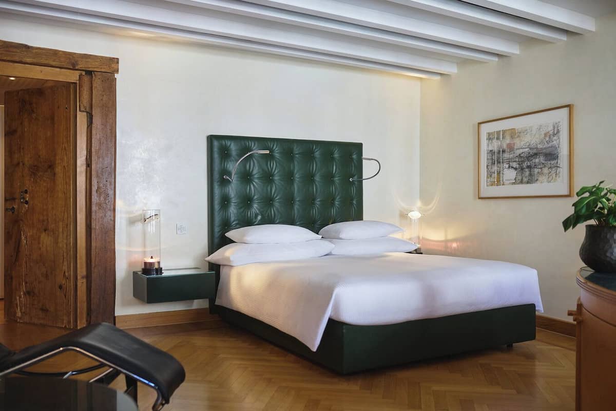 A neatly arranged bedroom with a green upholstered headboard, a bed with white linens, a bedside table with a lamp, and a framed artwork on the wall.
