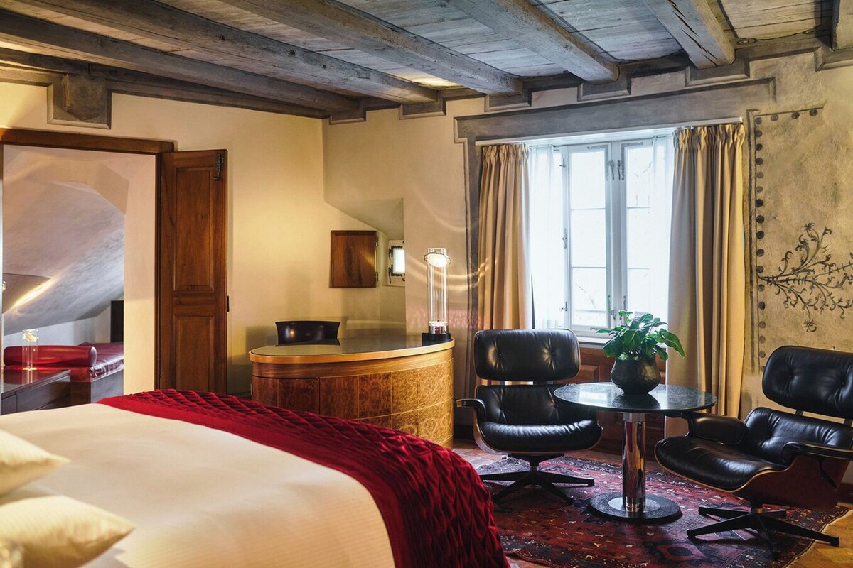 Ein Hotelzimmer mit Bett, Schreibtisch, zwei Stühlen, einem kleinen Tisch und einem Fenster mit Vorhängen.