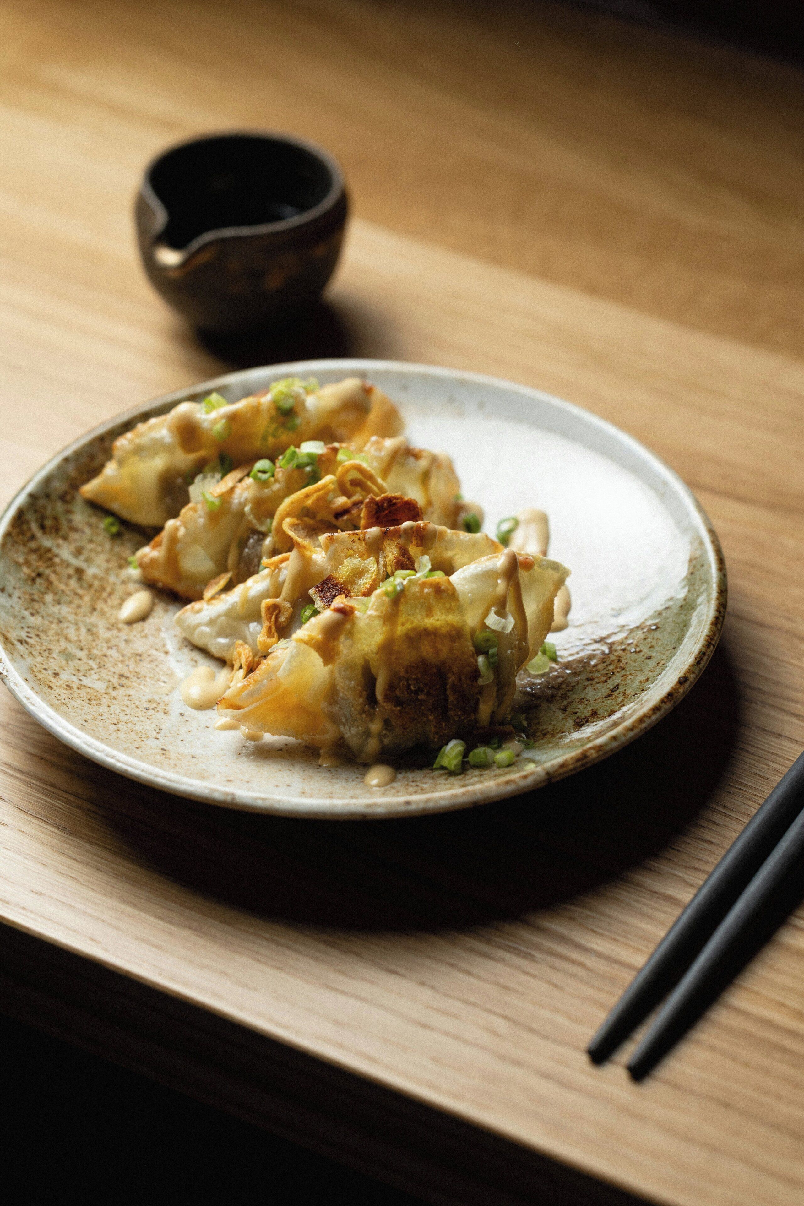 A plate of dumplings garnished with green onions and a small bowl of sauce, placed on a wooden table.
