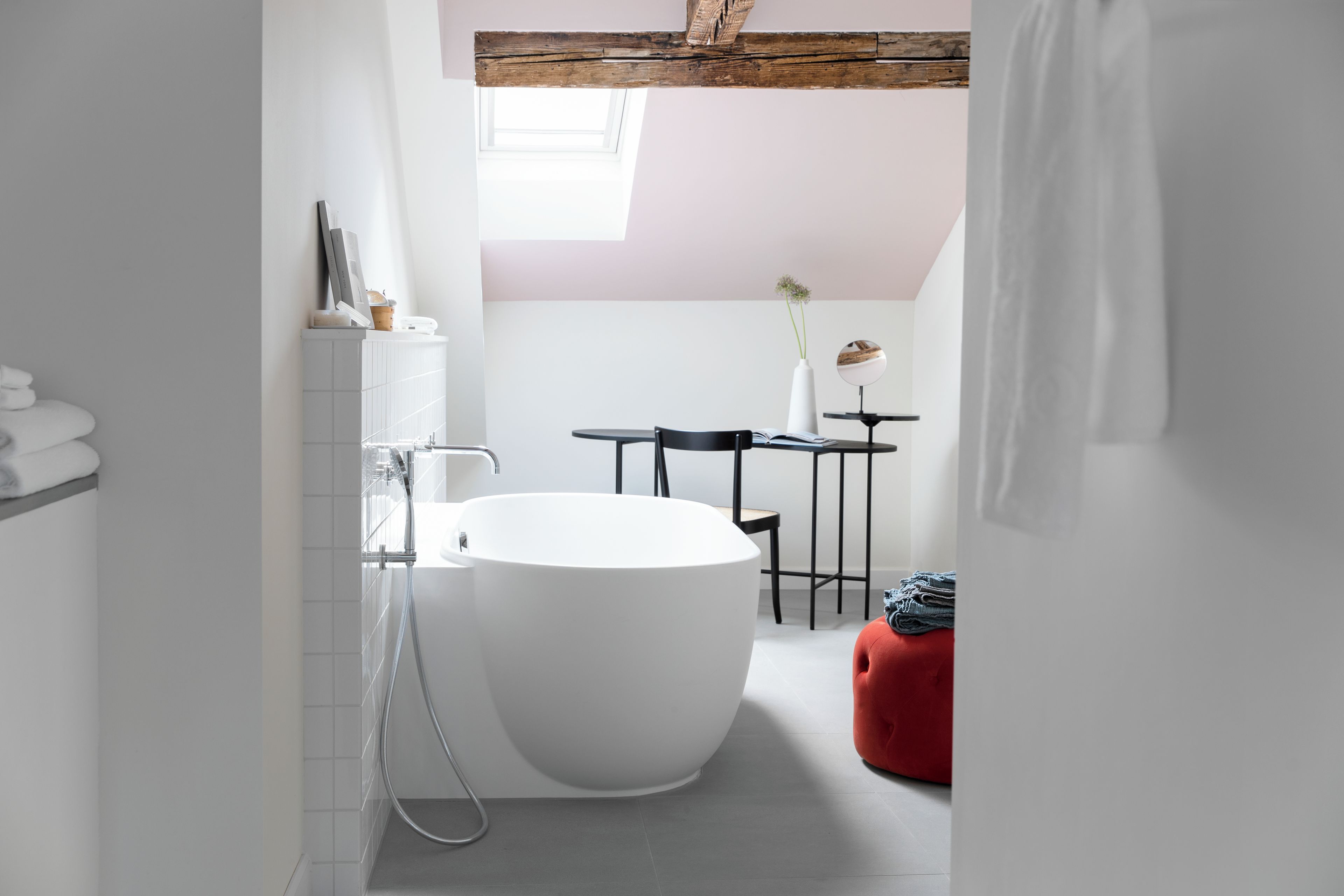 A modern bathroom with a freestanding bathtub, a small table with a chair, and a red ottoman.