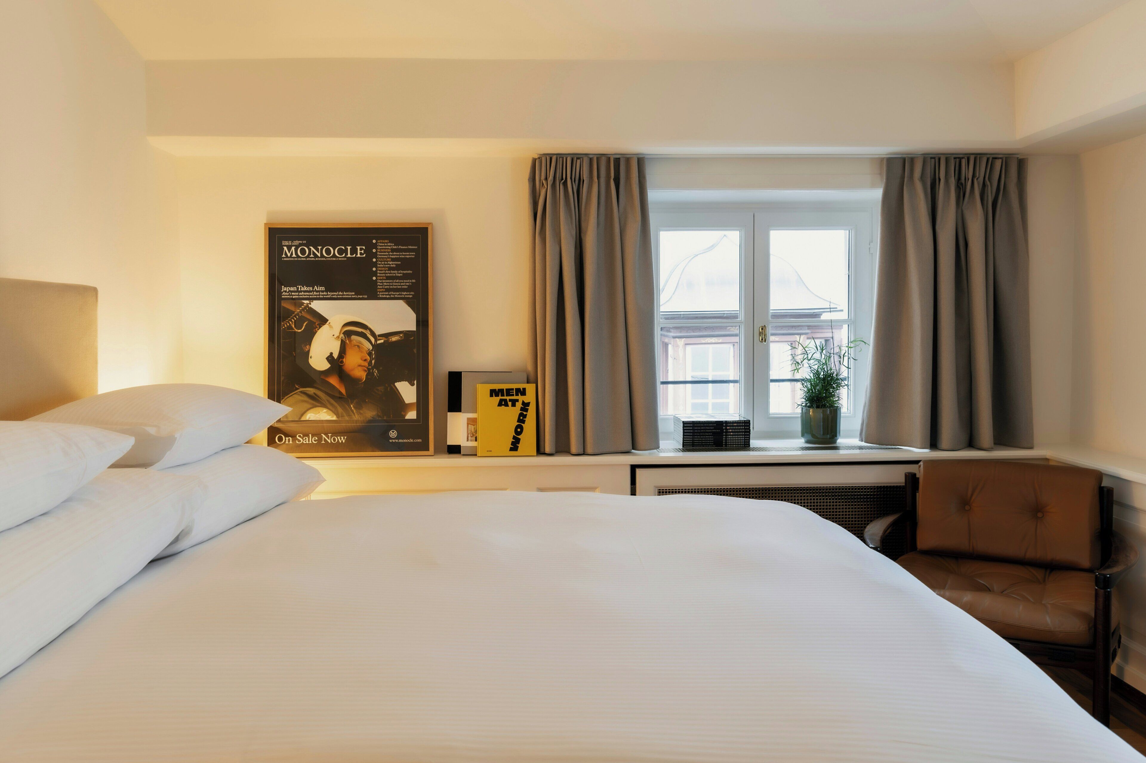 Habitación de hotel con una cama grande, una silla, un ventanal con cortinas, un cartel en la pared y una mesita con una planta.