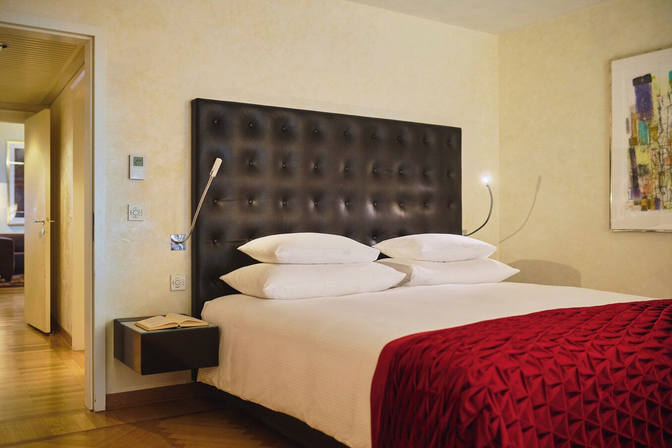 A hotel room with a large bed, a dark headboard, and a red blanket. There is a nightstand with a lamp and a small table with a book.