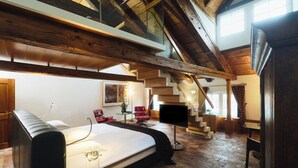 A spacious room with a large bed, a desk, a chair, a TV, and a staircase leading to an upper level. - Widder Hotel (Zürich)