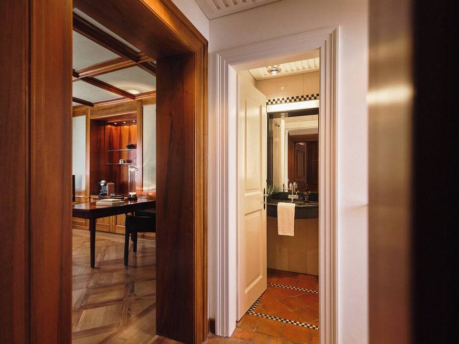 A hallway with a wooden door leading to a dining area with a table and chairs, and a kitchen area with a sink and countertop visible through an open door.
