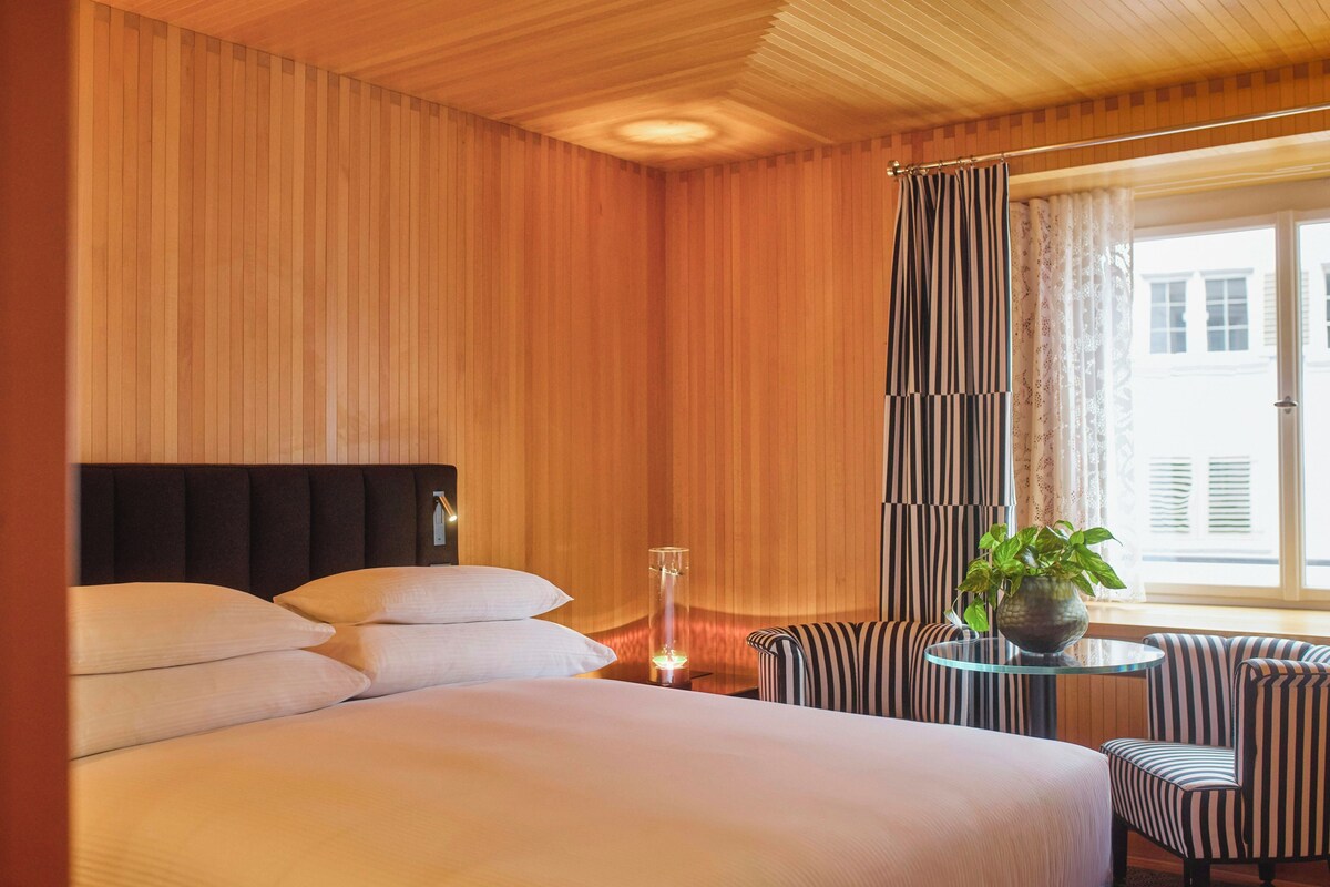 Ein Hotelzimmer mit einem großen Bett, einem Nachttisch mit Lampe, einem Sessel und einem Fenster mit Vorhängen.