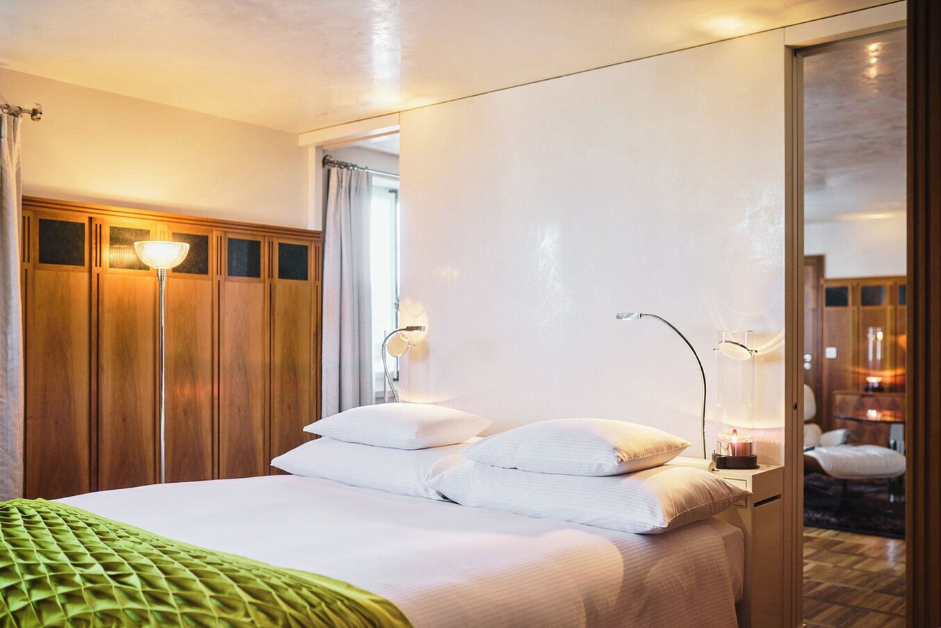 Ein Hotelzimmer mit einem Bett, Nachttischlampen, einer grünen Tagesdecke und Blick in den Innenraum durch eine Glastür.