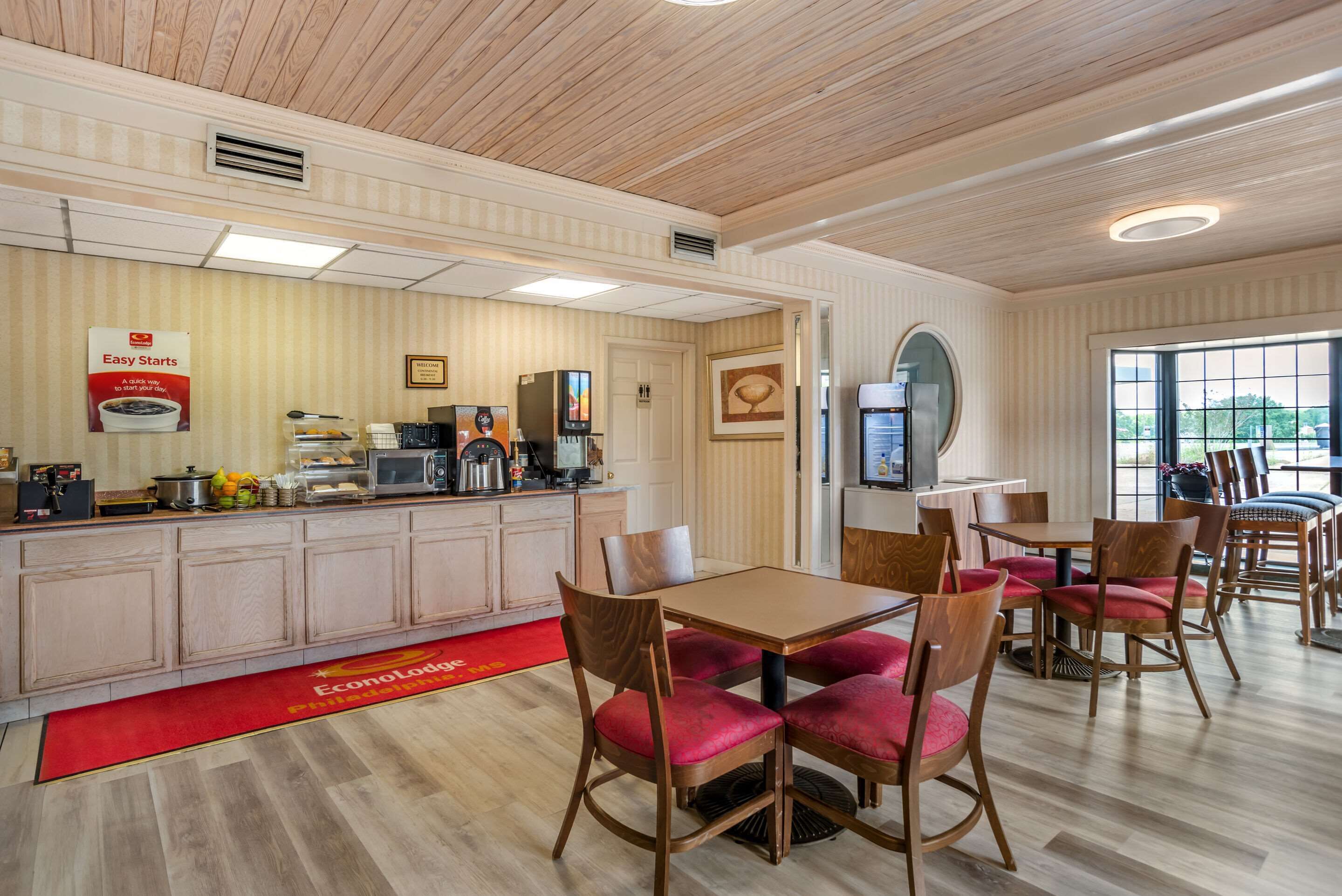 Un restaurante con techo de madera, una zona de cocina y un comedor con sillas tapizadas en rojo.