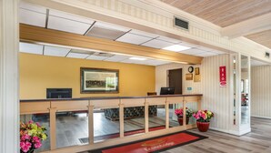 Uma área de recepção com balcão de madeira, um quadro na parede, um relógio e um tapete vermelho com o nome do hotel.