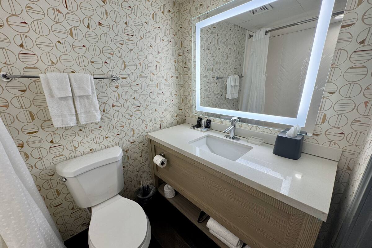 A bathroom with a toilet, sink, and mirror. There are towels hanging on a rack and a trash can.
