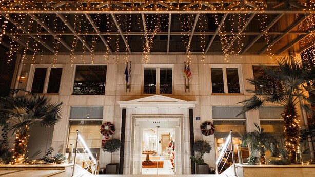 Uma entrada de edifício com galeria coberta decorada com luzes de cordão, palmeiras e guirlandas.