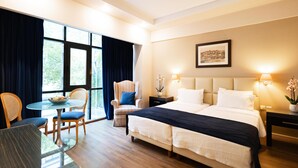 Quarto de hotel com uma cama grande, uma mesa de jantar com cadeiras, uma mesa pequena com uma tigela de frutas e uma janela com cortinas.