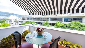 Uma varanda com toldo listrado, mesa de vidro, plantas em vasos e vista para prédios e vegetação.