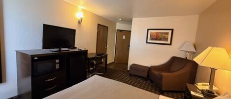 A hotel room with a bed, a desk with a television, a chair, a lamp, and a framed picture on the wall.