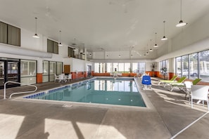 An indoor swimming pool with lounge chairs and tables, large windows, and ceiling lights.