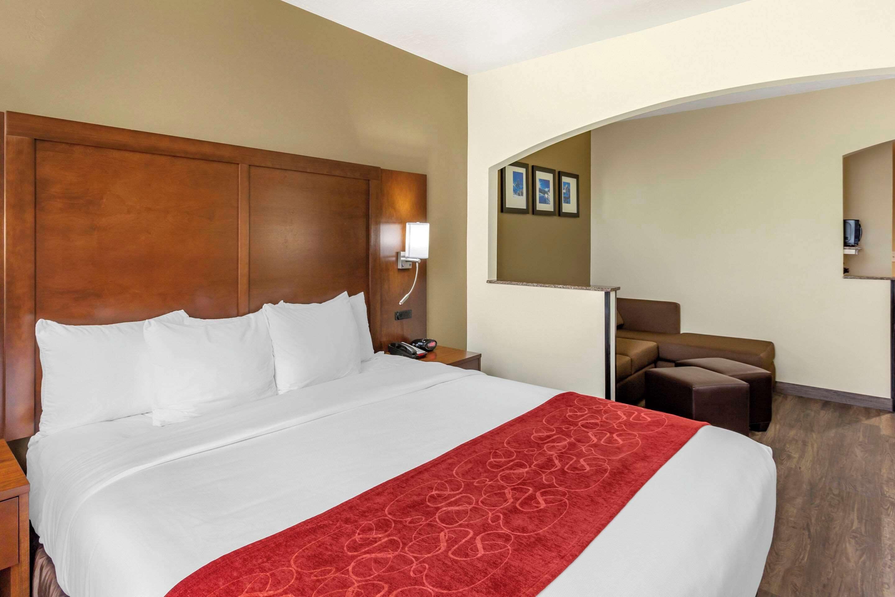 A hotel room with a large bed, a wooden headboard, a red throw blanket, a nightstand with a phone, and a seating area with a couch and armchair.