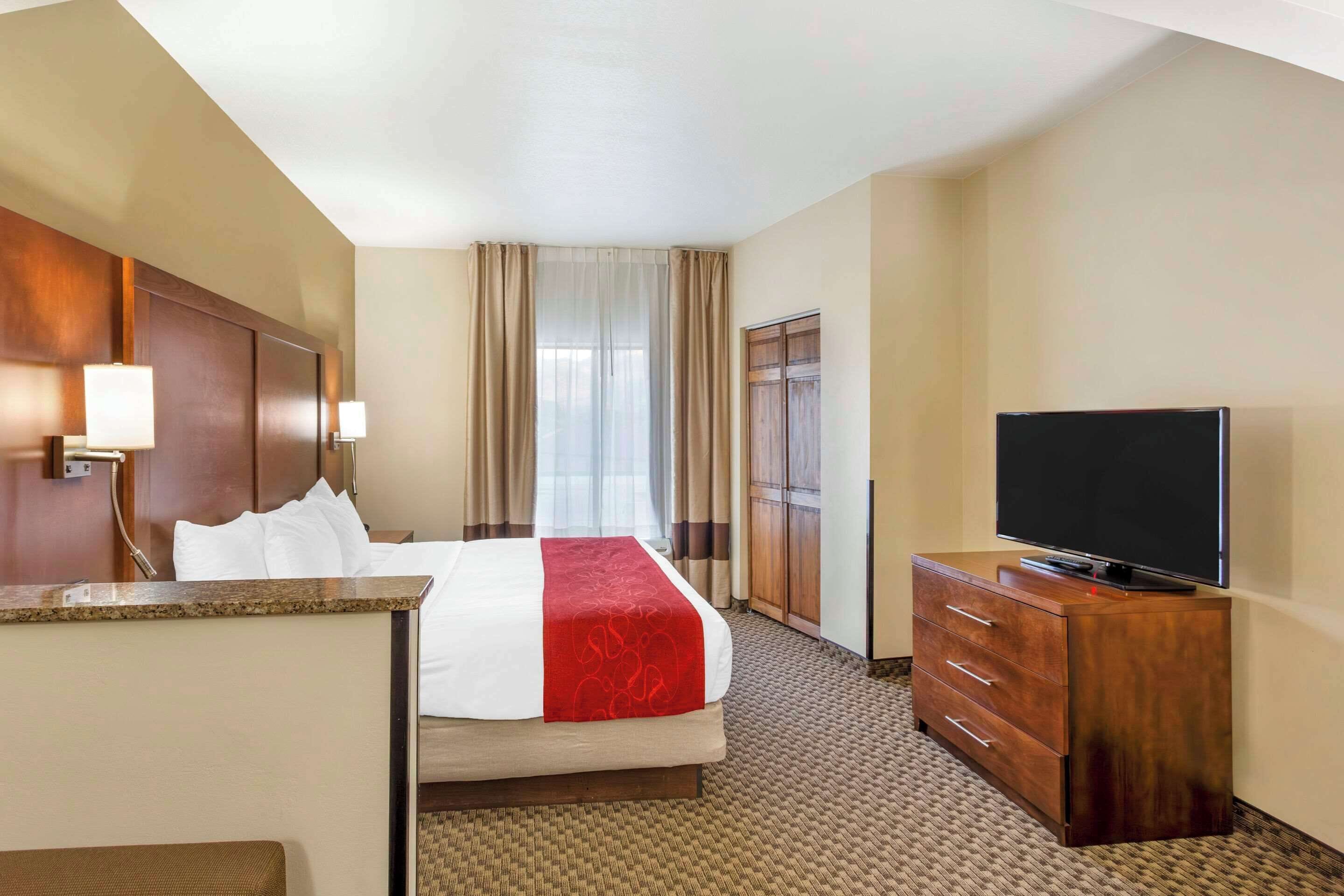A hotel room with a bed, a TV on a wooden dresser, a desk with a lamp, and a window with curtains.
