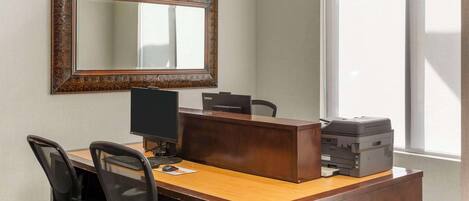 Una oficina de hotel con un escritorio de madera, dos sillas de oficina negras, un ordenador de sobremesa, una impresora y un espejo grande en la pared.
