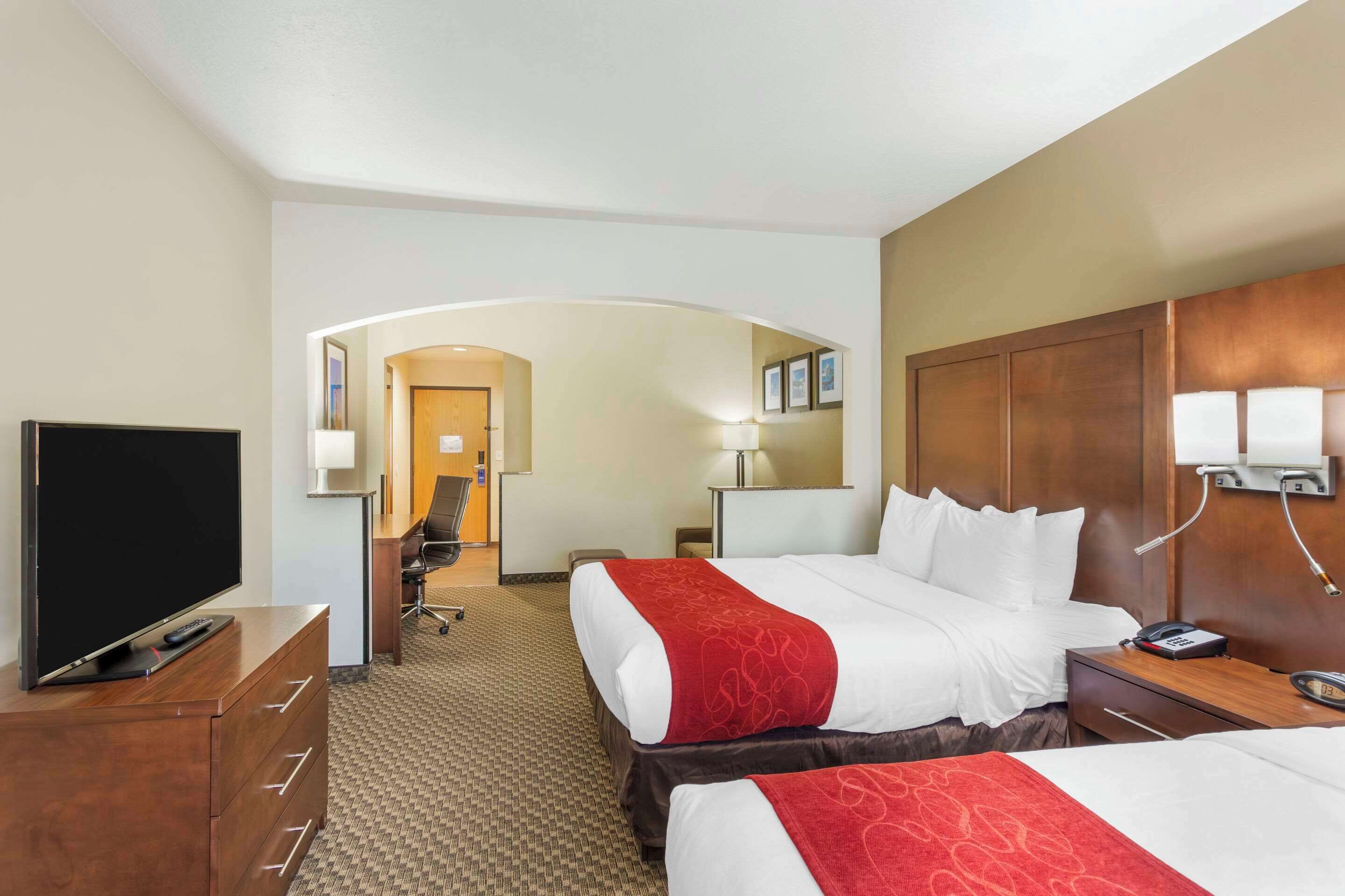 A hotel room with two beds, a television on a wooden dresser, a desk with a lamp, and a hallway leading to another room.