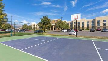 An outdoor tennis court with a basketball hoop, surrounded by a fence, and adjacent to a parking lot with several cars.