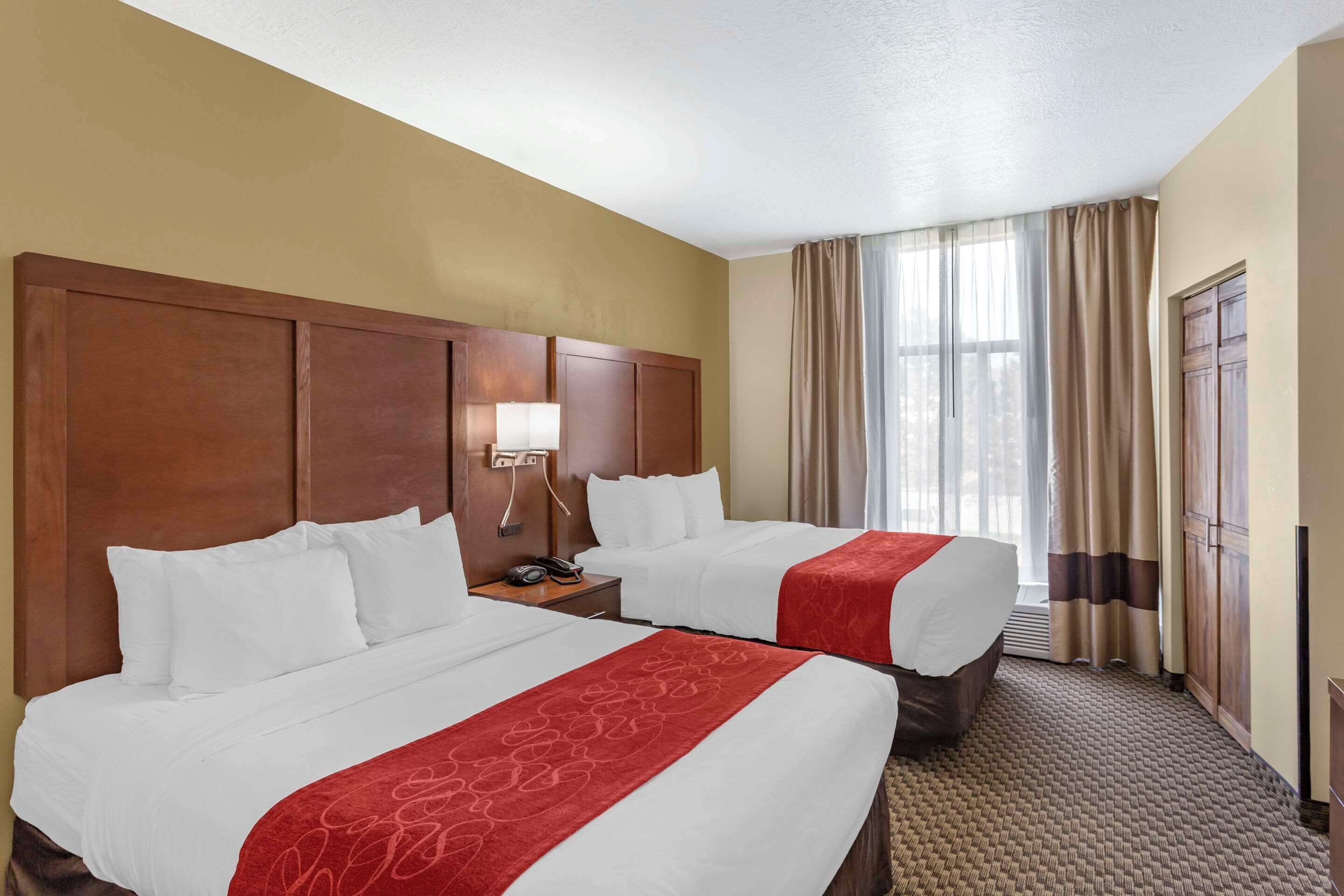 A hotel room with two beds, a wooden headboard, a nightstand with a lamp, and a large window with curtains.