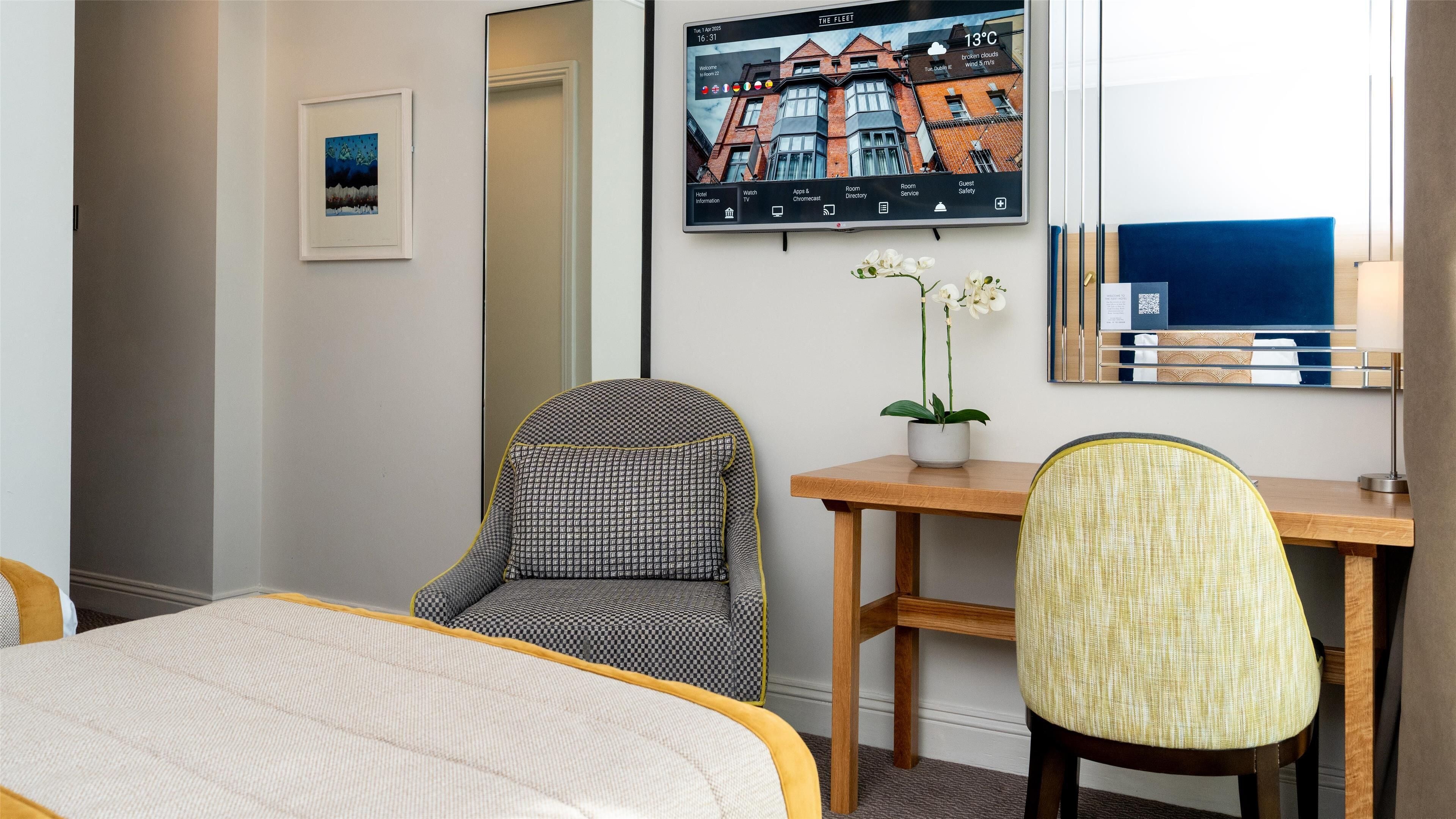 A hotel room with a bed, two chairs, a desk, a TV, and a framed picture of a building.