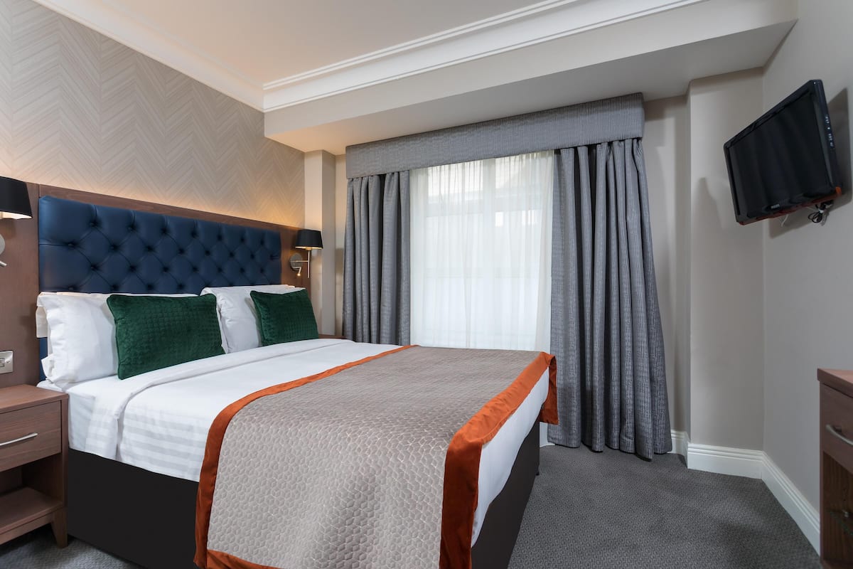 Una habitación de hotel con una cama grande, cabecera, mesita de noche, televisor montado en la pared y ventana con cortinas.