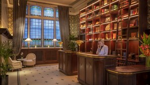 Een receptiezone met een houten balie, twee stoelen, een grote boekenkast en een raam met glas-in-lood.