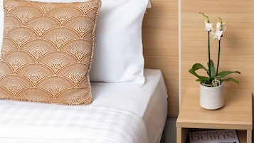 A neatly made bed with white and patterned pillows, a small potted orchid on a nightstand, and a magazine titled "DINING IN DUBLIN" under the nightstand.