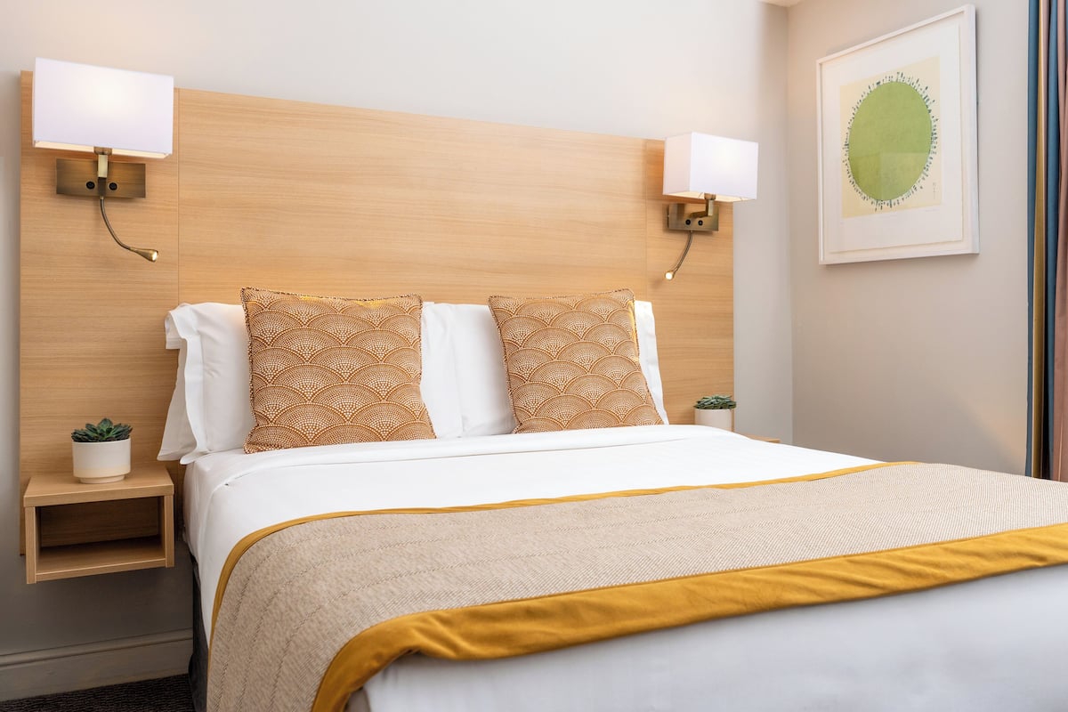 Habitación de hotel con una cama grande, dos mesitas de noche, una lámpara de pared, una obra de arte enmarcada y una ventana con cortinas.