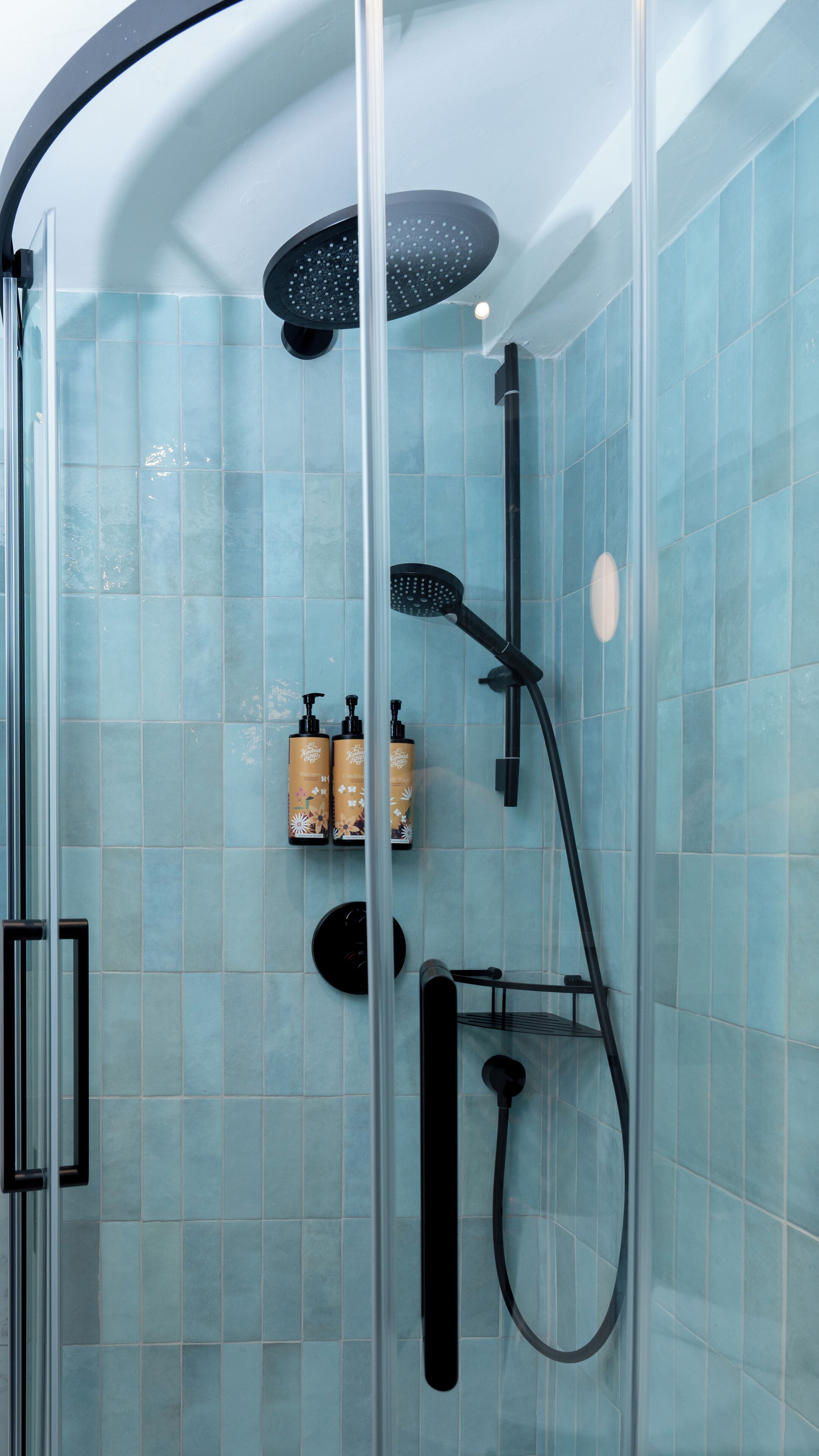 A glass-enclosed shower with a handheld showerhead and a wall-mounted rain showerhead. There are two bottles of shampoo and conditioner on a shelf.