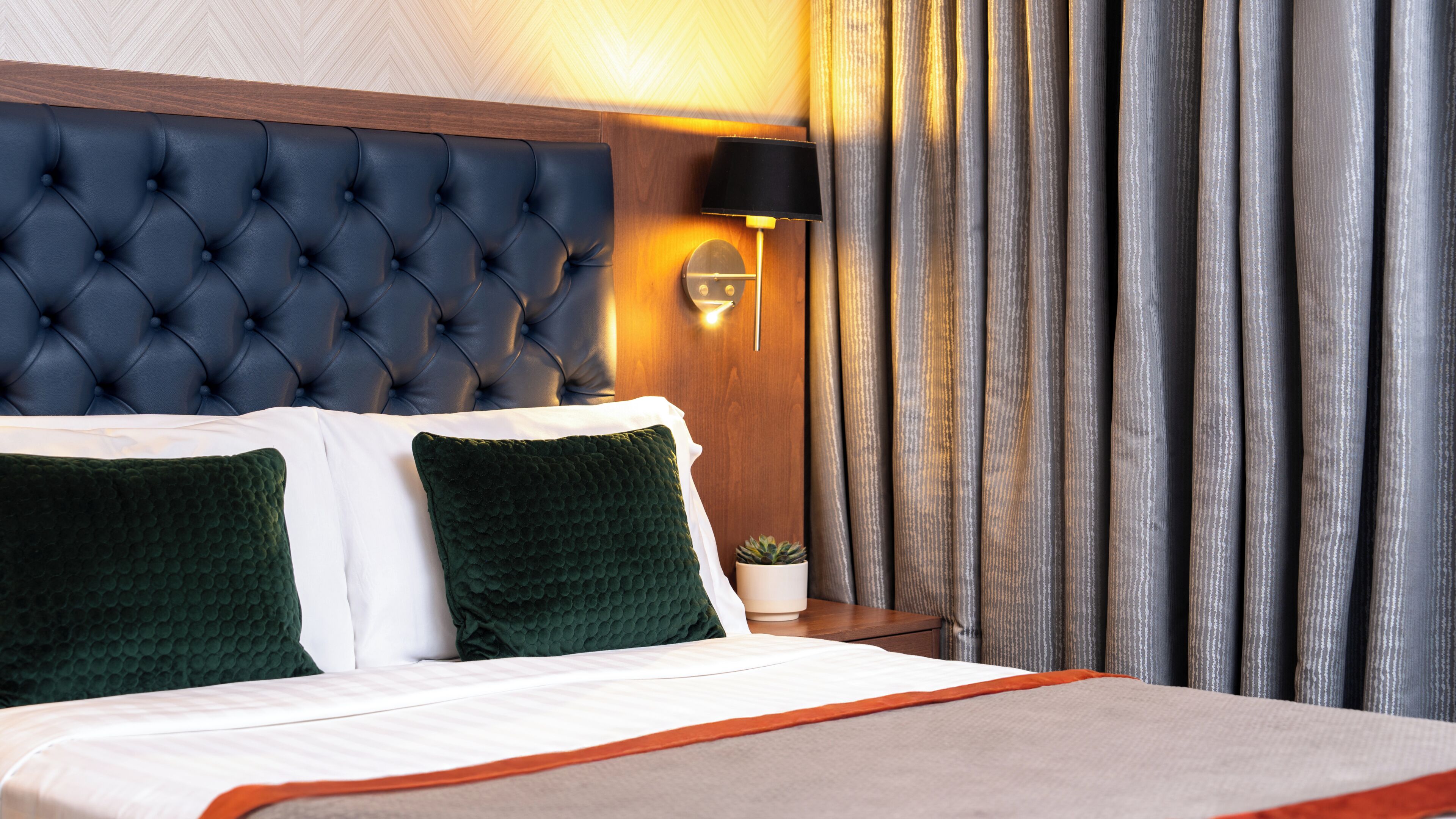 Hotellihuone, jossa on koristeltu päätyseinä, valkoisilla ja vihreillä tekstiileillä varustettu sänky, yöpöytälamppu ja verho.