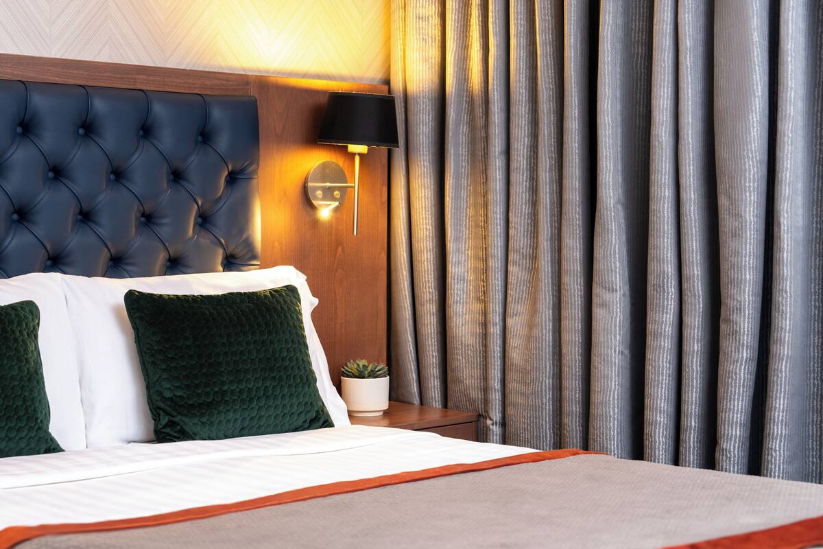A hotel room with a tufted headboard, a bed with white and green bedding, a bedside lamp, and a curtain.