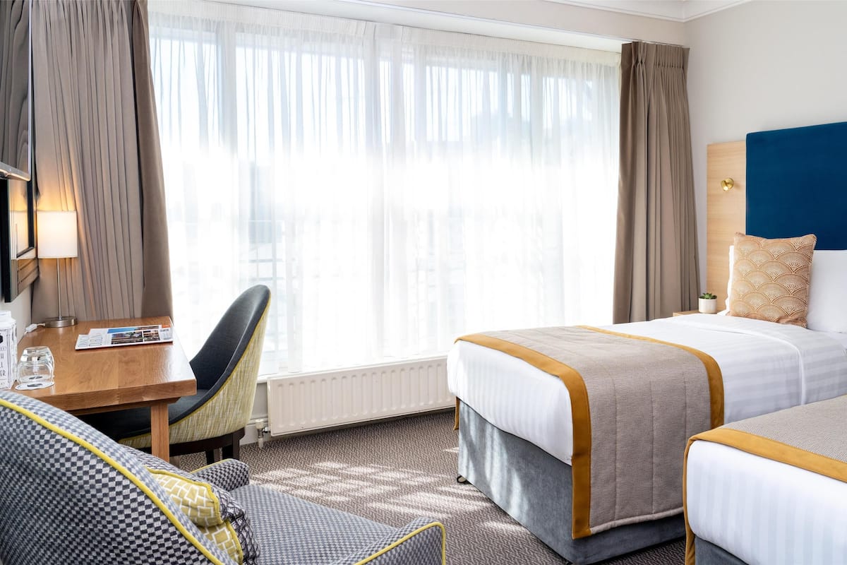 Habitación de hotel con dos camas, un escritorio, una silla y un ventanal con cortinas.
