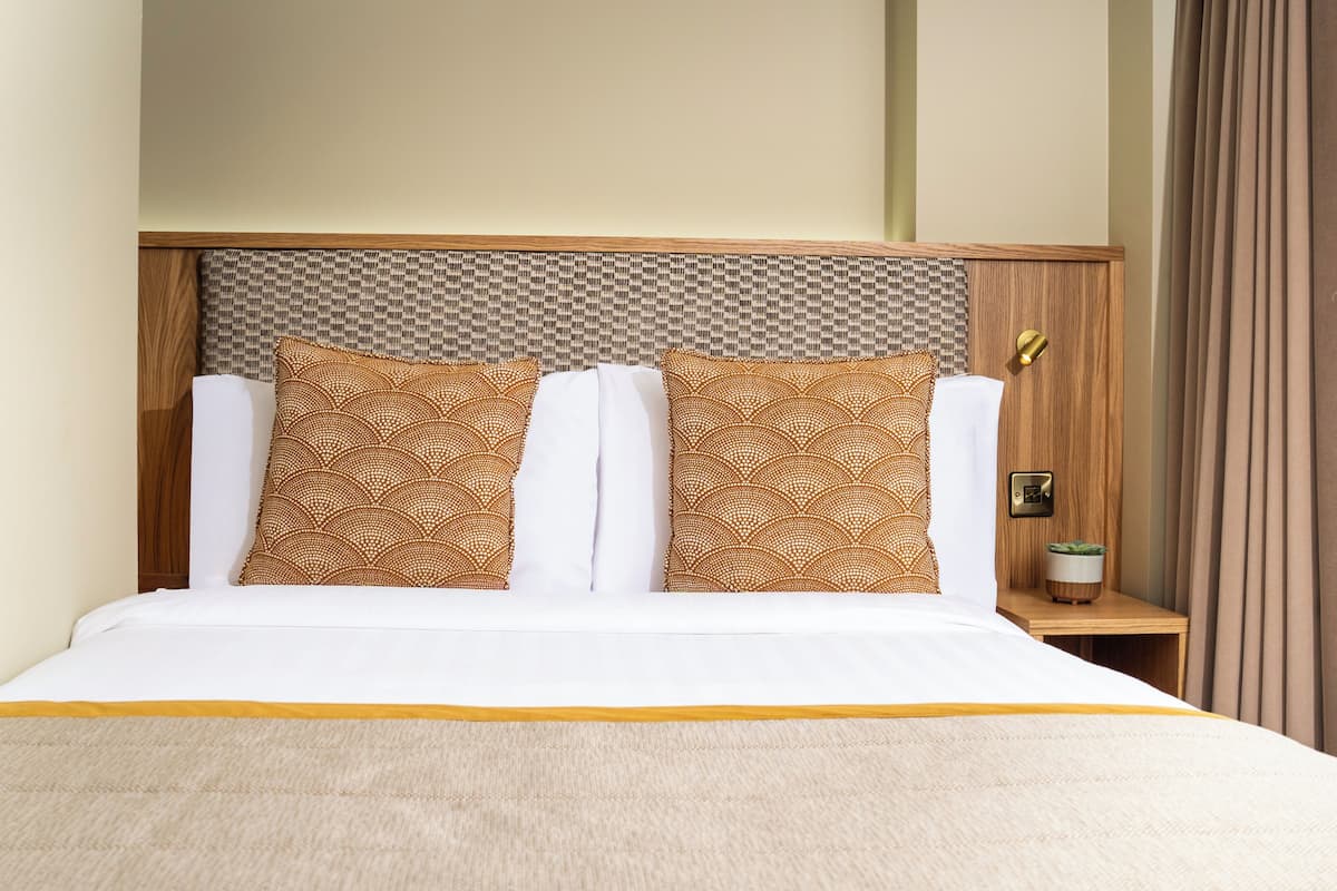 Habitación de hotel con cama, dos almohadas estampadas, una mesita de noche con lámpara y ventana con cortinas.
