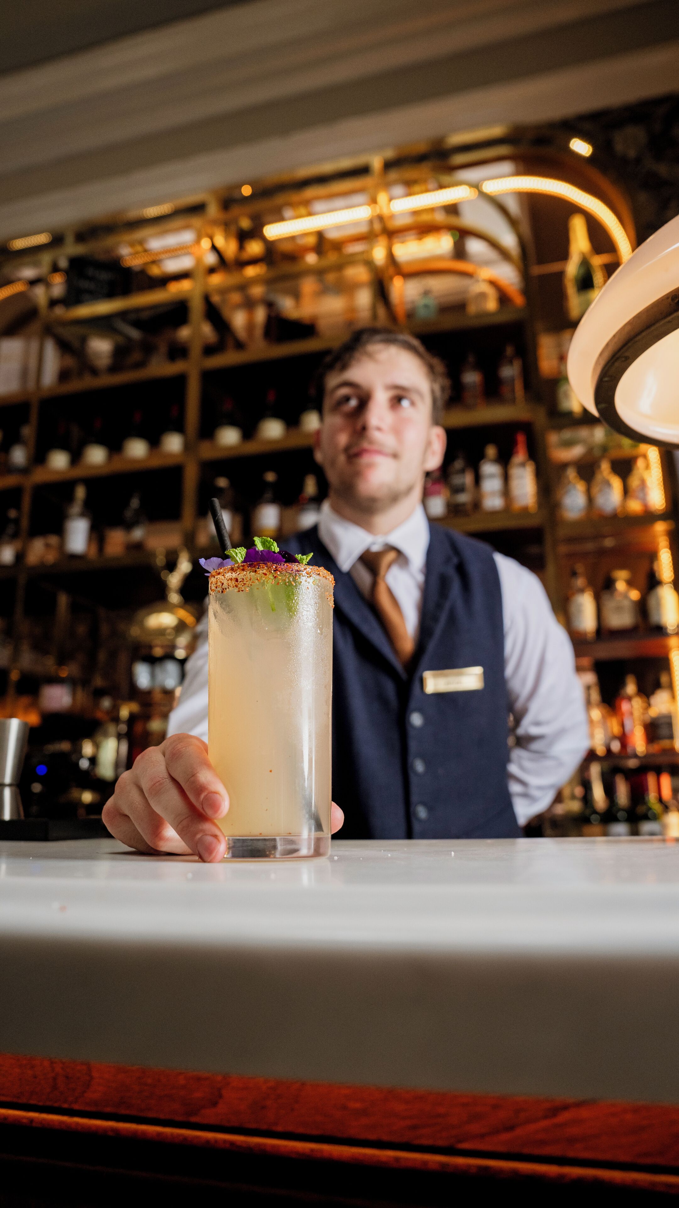 Un cantinero sosteniendo un cóctel con decoración, en un bar con estantes llenos de botellas al fondo.